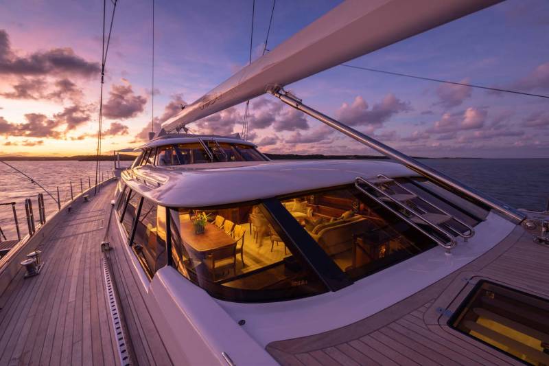 A luxurious yacht's deck stretches into the foreground, with a warmly lit interior visible through its windows, while a colorful sunset paints the sky over a calm ocean in the background.