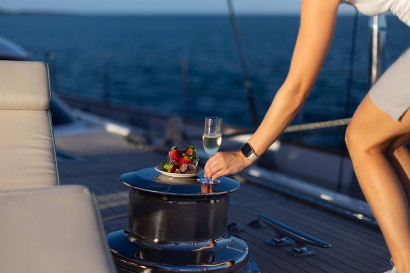 A woman's arm reaches for a plate of strawberries and a glass of champagne on a yacht's deck, with the calm blue ocean stretching out in the background.