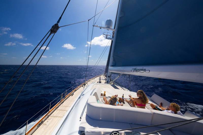 People relax on the deck of a sailboat with a blue sail against a bright blue sky and ocean.