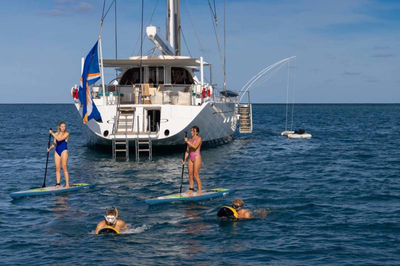 Two women paddleboard and two people snorkel in the foreground, with a large white yacht anchored in the blue ocean behind them.