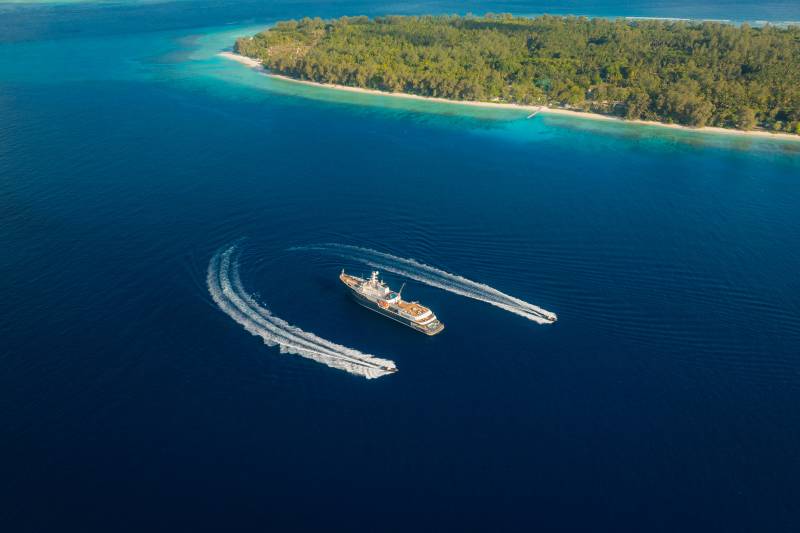 A large yacht cruises through deep blue water, trailed by two smaller boats, with a lush green island in the background.