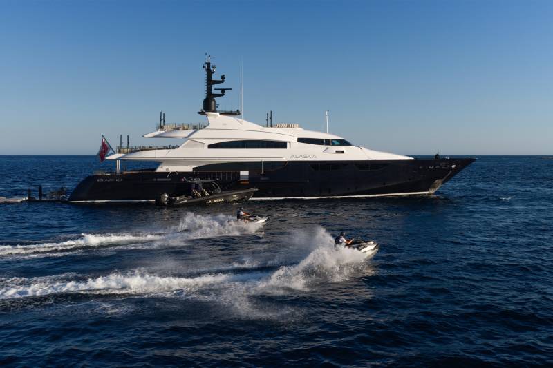 Two jet skis create white wakes in the foreground of a large, dark-hulled luxury yacht named "Alaska" under a clear blue sky.