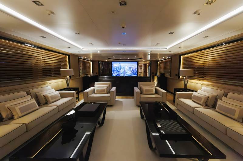 Two long white sofas and black coffee tables face each other in the foreground of a luxurious lounge, with a large screen displaying a city skyline in the background.