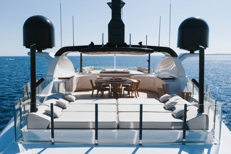 A luxury yacht deck features white lounge seating and a wooden dining area in the foreground, set against a vast blue ocean and clear sky in the background.
