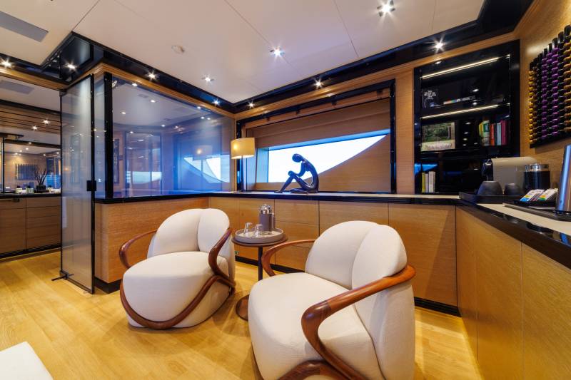 Two white curved armchairs sit on a light wood floor in the foreground, with a modern lounge area featuring a wine rack and a window view in the background.