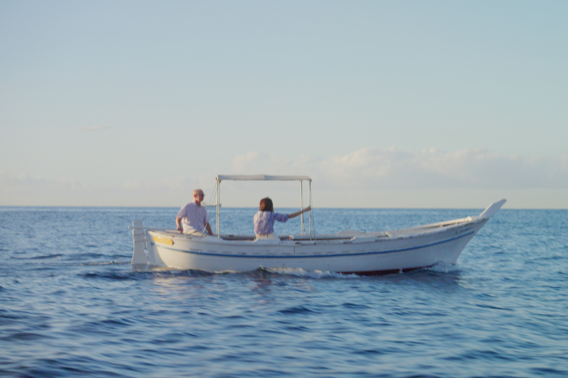 Two people ride in a small white boat across a calm blue ocean under a clear sky.
