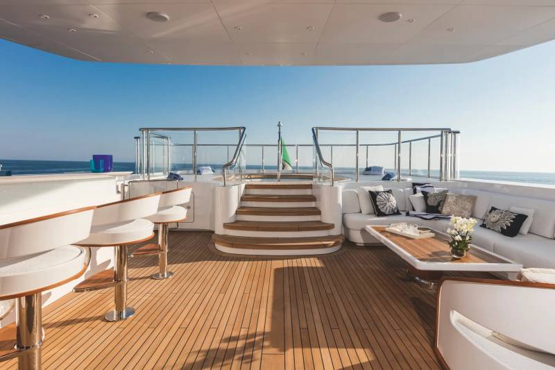 A luxury yacht deck features white seating and a wooden floor in the foreground, leading to stairs and a railing with a clear blue ocean and sky in the background.