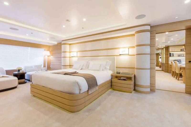 A luxurious bedroom features a large bed with white linens in the foreground, set against a backdrop of cream-colored paneled walls and an open doorway leading to another room.