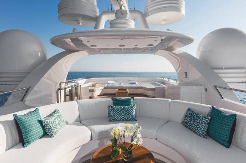 A white curved outdoor sofa with teal pillows and a flower arrangement sits in the foreground of a luxury yacht deck overlooking a calm blue ocean.