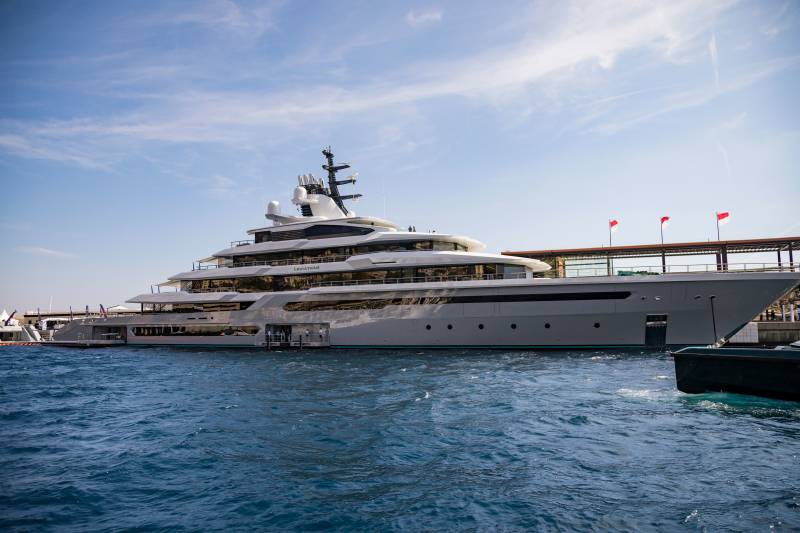 A massive luxury yacht floats on deep blue water against a backdrop of a clear sky and a distant pier with red flags.