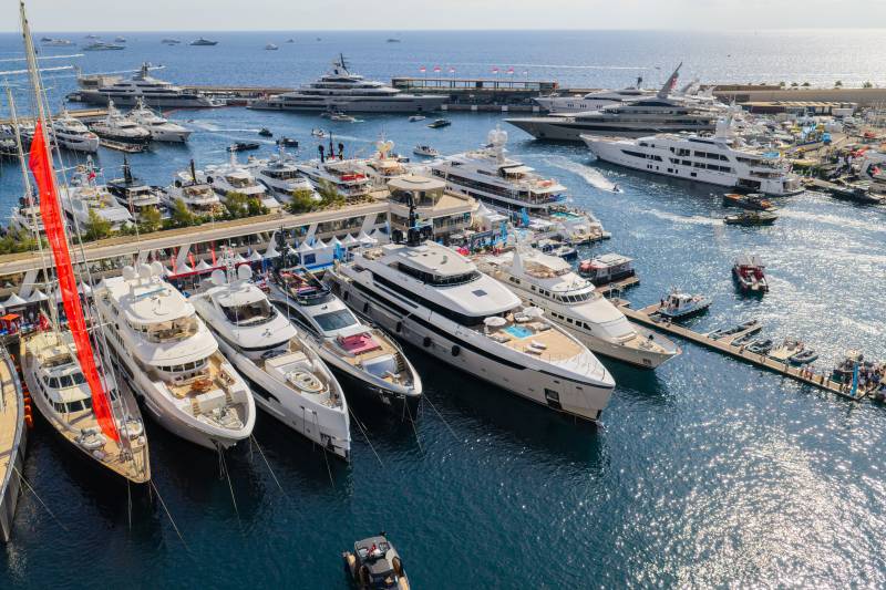 Numerous luxury yachts are docked in a crowded harbor, with a calm blue sea and a distant horizon in the background.