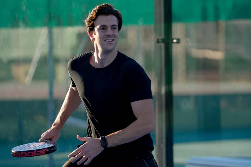 A man in a black t-shirt holds a padel racket in the foreground against a blurred background of a sports court and green fencing.