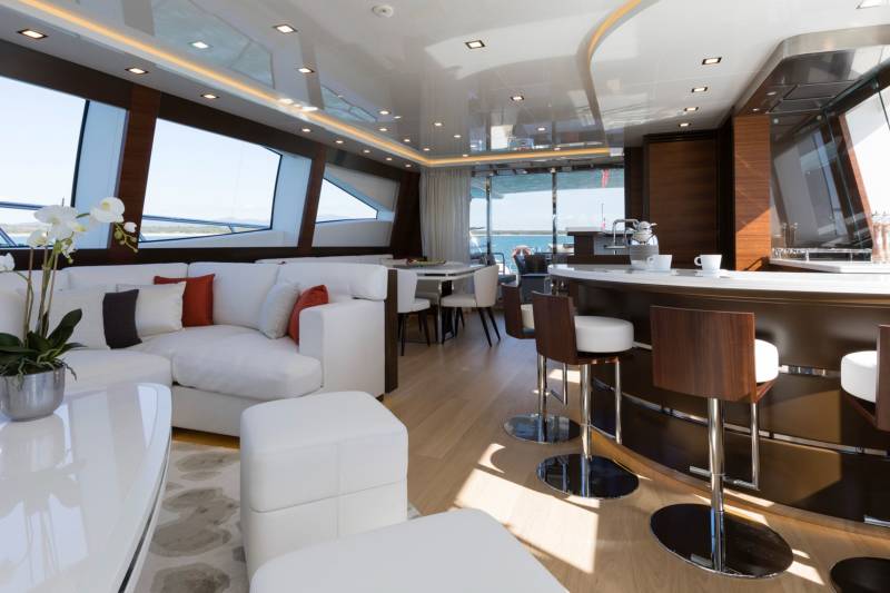 A modern yacht interior features a white sectional sofa and coffee table in the foreground, with a sleek breakfast bar and dining area set against large windows in the background.