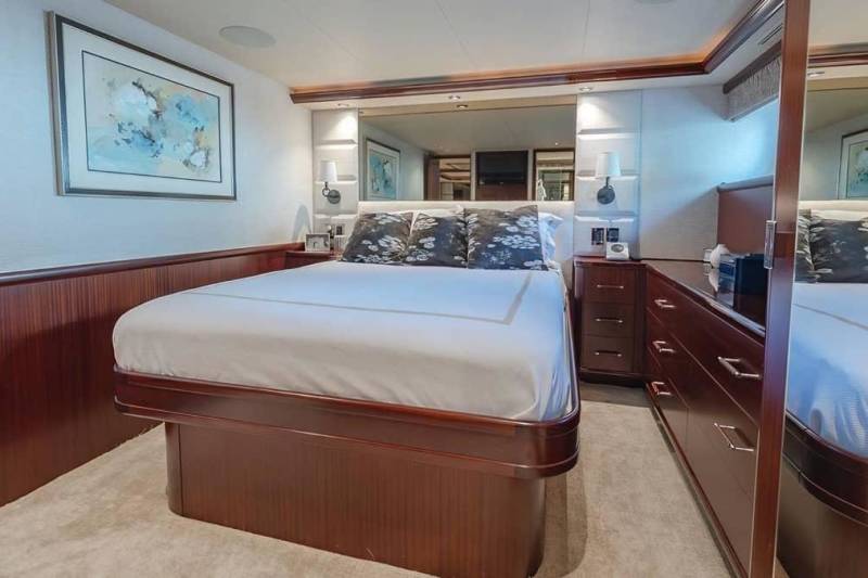 A neatly made bed with white linens and patterned pillows sits in the foreground of a luxurious bedroom featuring dark wood paneling and a large mirror in the background.