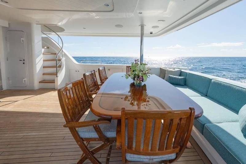 A wooden dining table and chairs sit on a yacht deck in the foreground, with a blue cushioned lounge and the open ocean in the background.