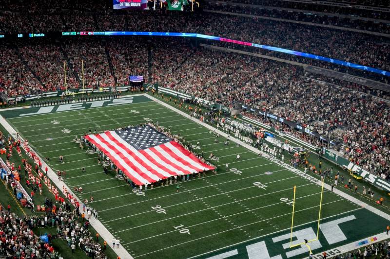 A massive American flag is spread across the center of a football field, surrounded by a crowd of spectators in a large stadium.
