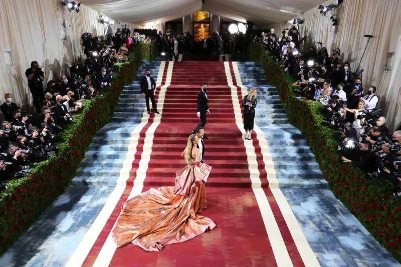 A woman in a voluminous copper gown walks up a red-carpeted staircase surrounded by crowds of photographers and guests.
