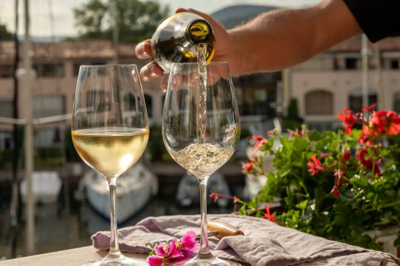 White wine is poured from a bottle into a glass next to another filled glass on a table, with red flowers and a blurred waterfront town in the background.