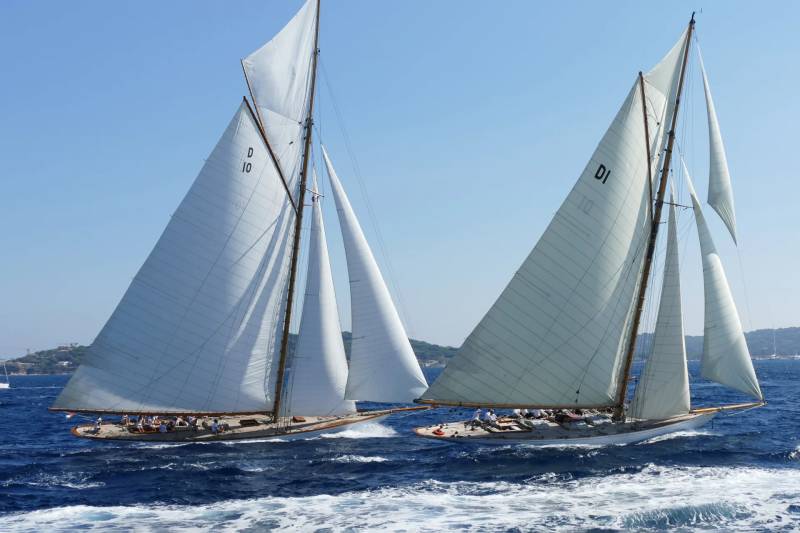 Two large white sailboats race across a choppy blue sea against a clear sky and distant coastline.