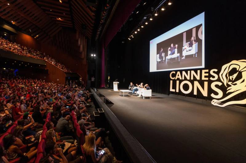 A large audience fills a theater, facing a stage where speakers are seated in front of a screen and a "Cannes Lions" sign.