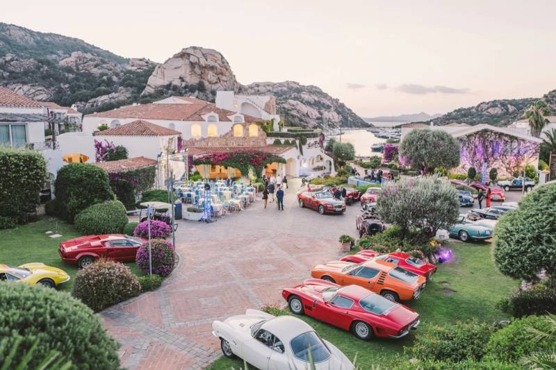 Luxury sports cars are parked on a paved courtyard in front of a Mediterranean villa, with rocky mountains and a harbor in the background.