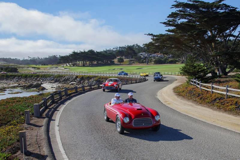 A line of vintage sports cars drives along a winding coastal road, with a lush green golf course and cypress trees in the background.