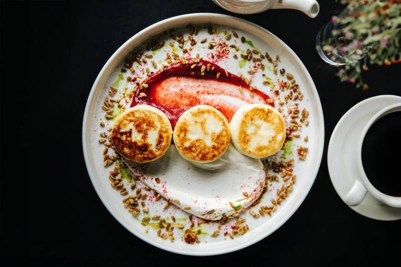 Three golden-brown cheese pancakes sit atop a swirl of white cream and red berry sauce on a white plate, set against a dark background with a cup of coffee.