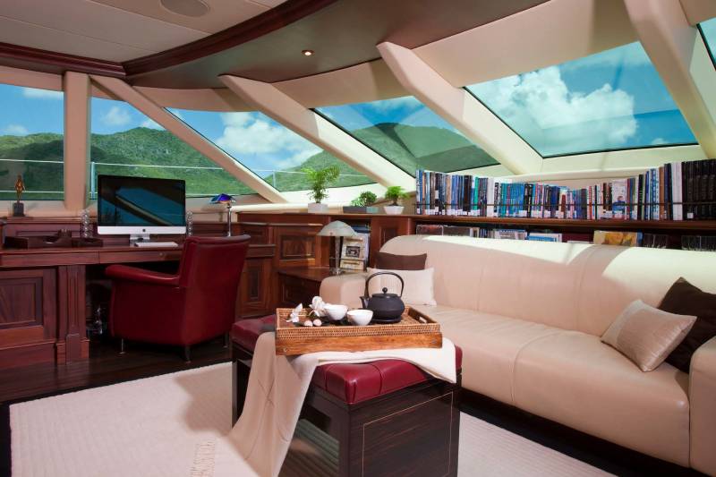 A luxurious lounge area with a white sofa and a red leather desk chair sits in the foreground, while large panoramic windows in the background reveal a view of green mountains under a blue sky.