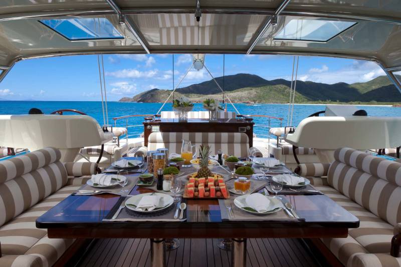 A set dining table with fruit and appetizers sits in the foreground of a luxury yacht, overlooking a turquoise ocean and lush green mountains in the background.