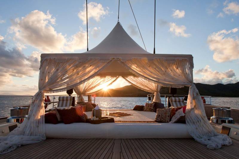 A luxurious white canopy bed with plush pillows sits on a wooden deck, set against a backdrop of calm ocean waters and distant mountains during a golden sunset.