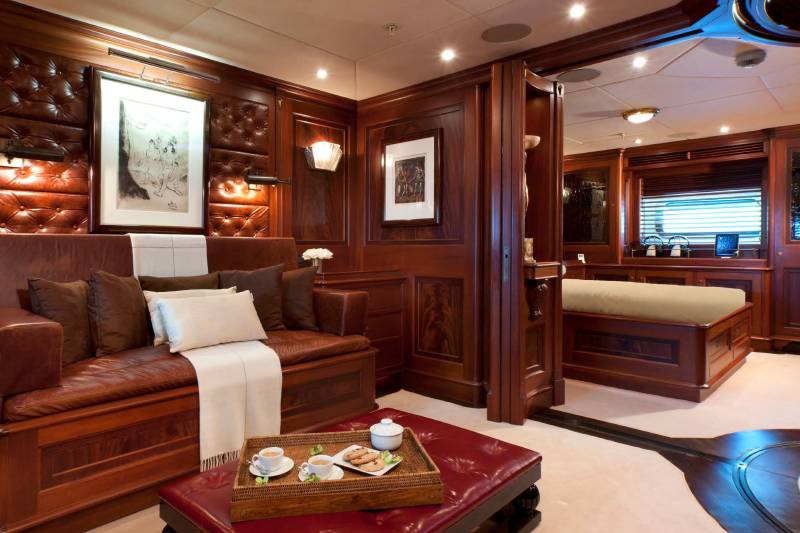 A leather sofa and a coffee tray sit in the foreground of a luxurious wood-paneled room, with a glimpse of a bedroom in the background.