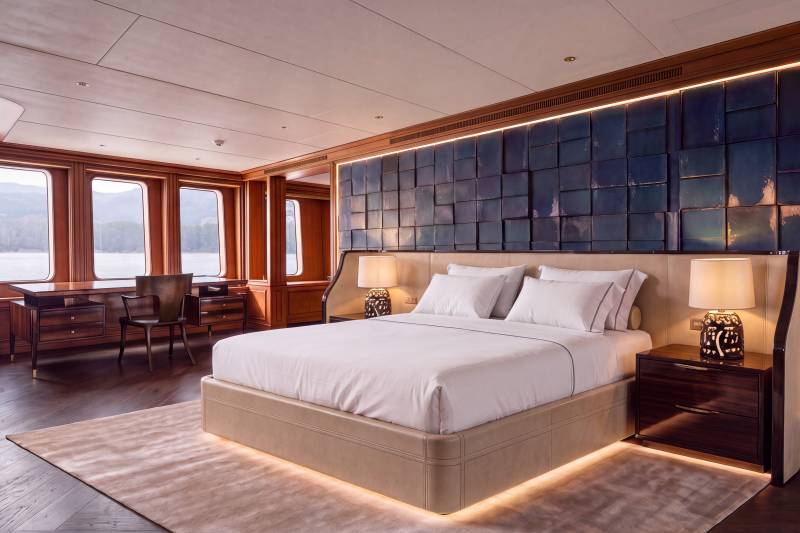 A luxurious bedroom features a large bed with white linens in the foreground, set against a backdrop of a textured blue wall and large windows overlooking a scenic waterfront.