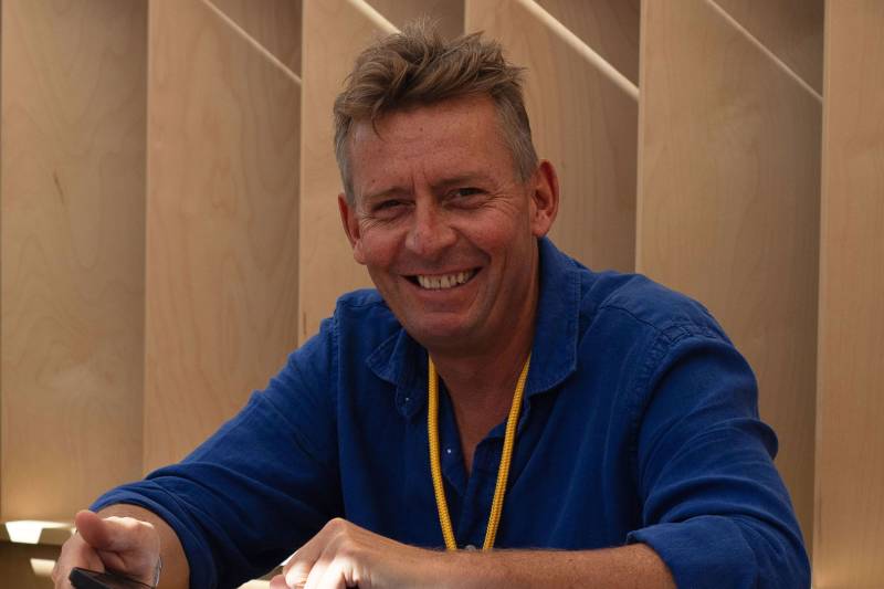 A smiling man in a blue shirt and yellow necklace sits in front of light-colored wooden shelving.