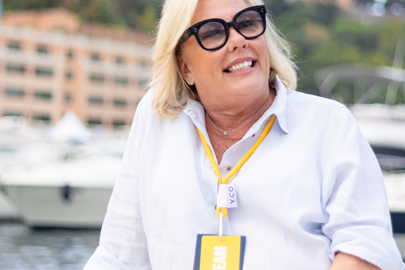 A smiling woman in a white shirt and sunglasses sits on a boat with a blurred cityscape and harbor in the background.