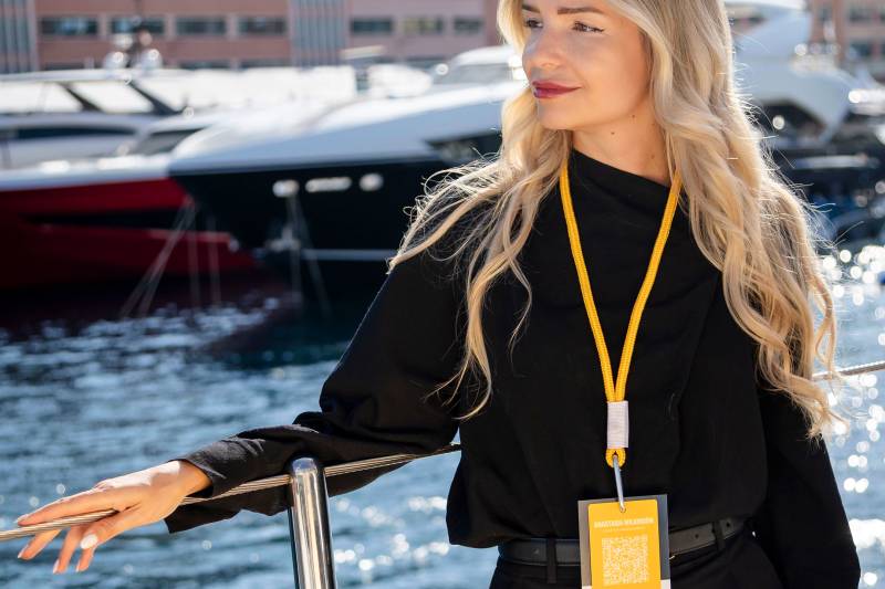 A blonde woman in a black outfit and yellow lanyard stands on a boat with yachts and coastal buildings in the background.