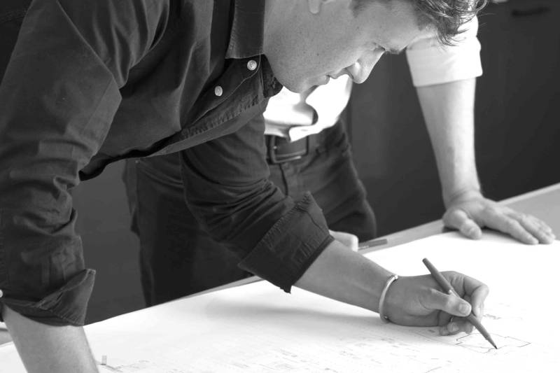 A man in a dark shirt leans over a table to draw on a large architectural plan while another person stands beside him in a blurred office setting.