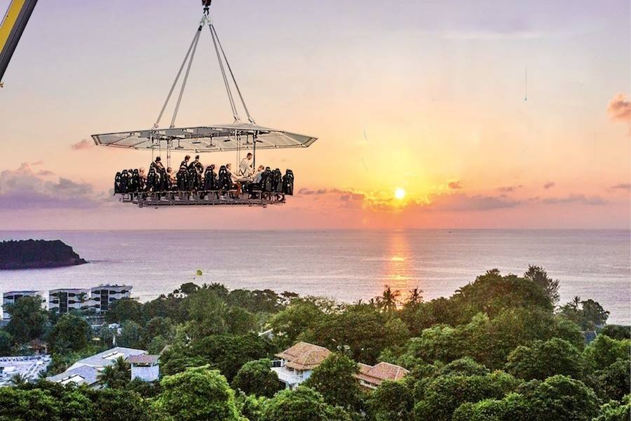 A suspended dining platform with people enjoying a meal hovers over a lush green landscape and a tranquil ocean at sunset.