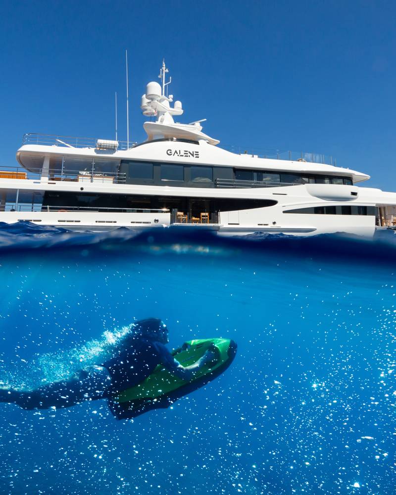 A diver explores the vibrant blue depths beneath a luxurious yacht sailing on a clear day.