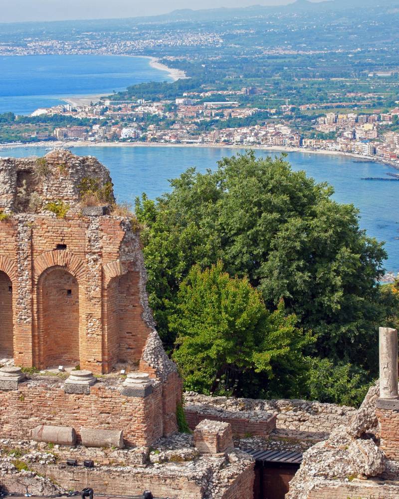 Ancient Roman ruins overlook a picturesque bay and coastal town under a clear blue sky.
