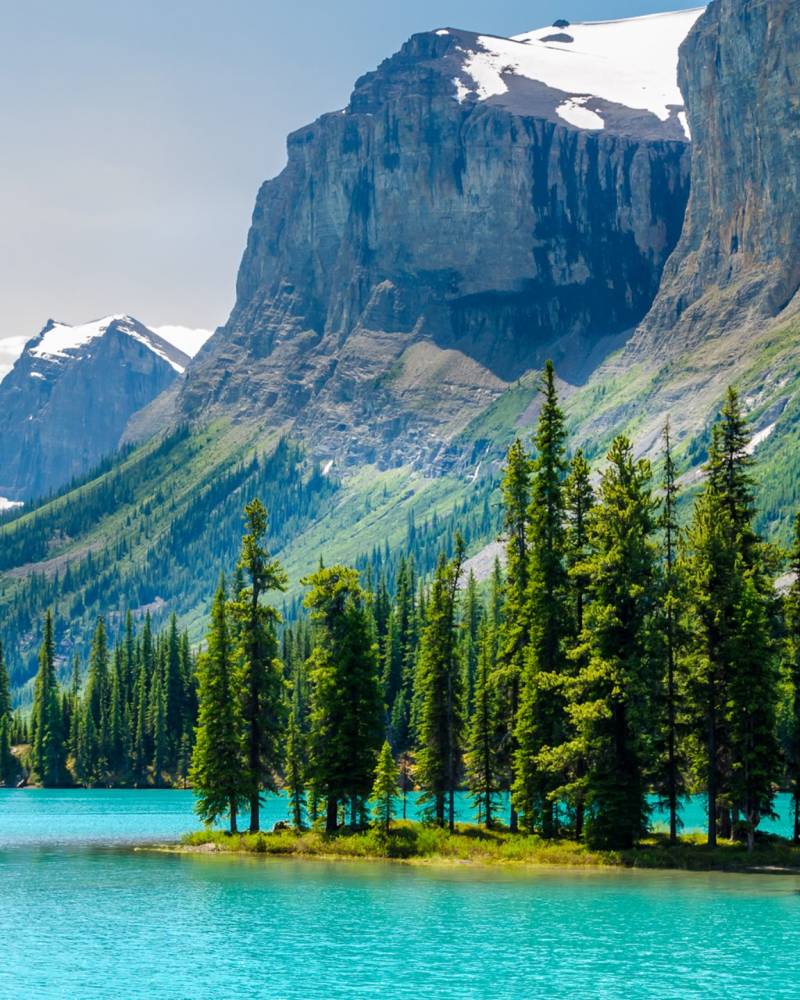 Turquoise waters surround a small island dotted with evergreen trees, set against a backdrop of majestic, snow-capped mountains.
