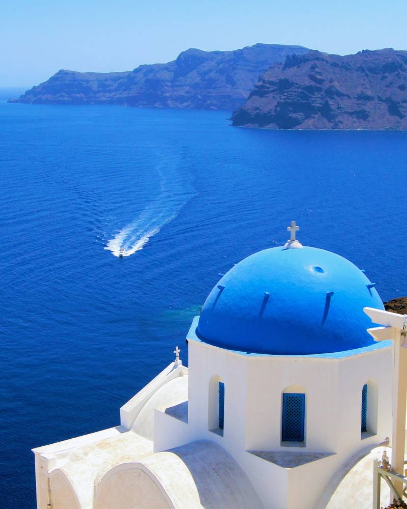 A classic blue-domed church overlooks the sparkling Aegean Sea with a boat leaving a white wake.