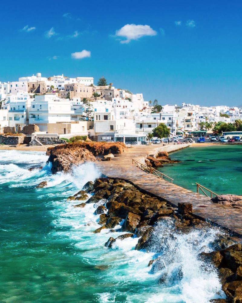 Waves crash against a stone pier leading to a charming white-washed village nestled on a hillside overlooking the turquoise sea.
