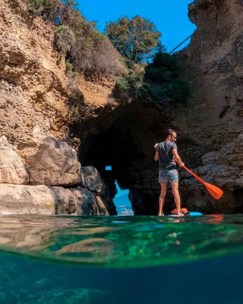 A paddleboarder explores a sea cave, with the clear turquoise water revealing the vibrant underwater world.