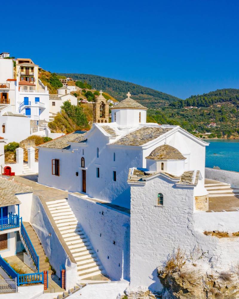 A whitewashed church with a stone-tiled roof perches dramatically on a cliff overlooking the turquoise Aegean Sea.