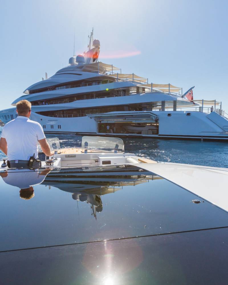 A man pilots a small boat towards a massive, multi-decked superyacht anchored in the azure sea.