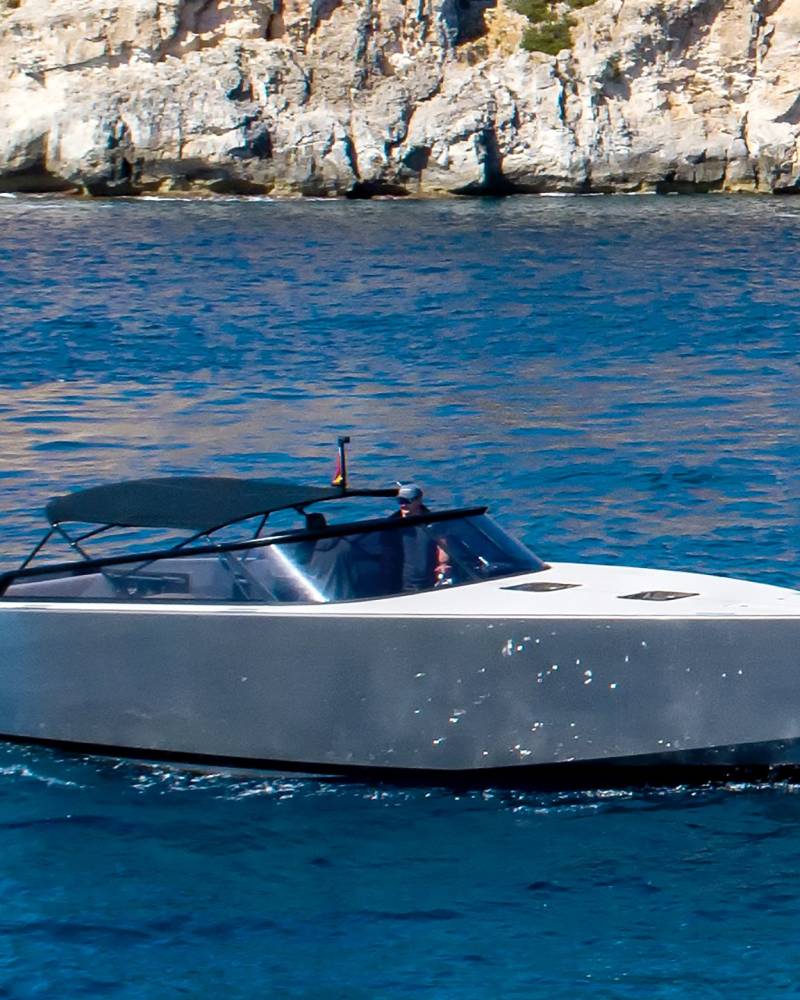 A sleek, modern yacht glides through the vibrant blue waters near a rugged, sun-drenched cliff face.