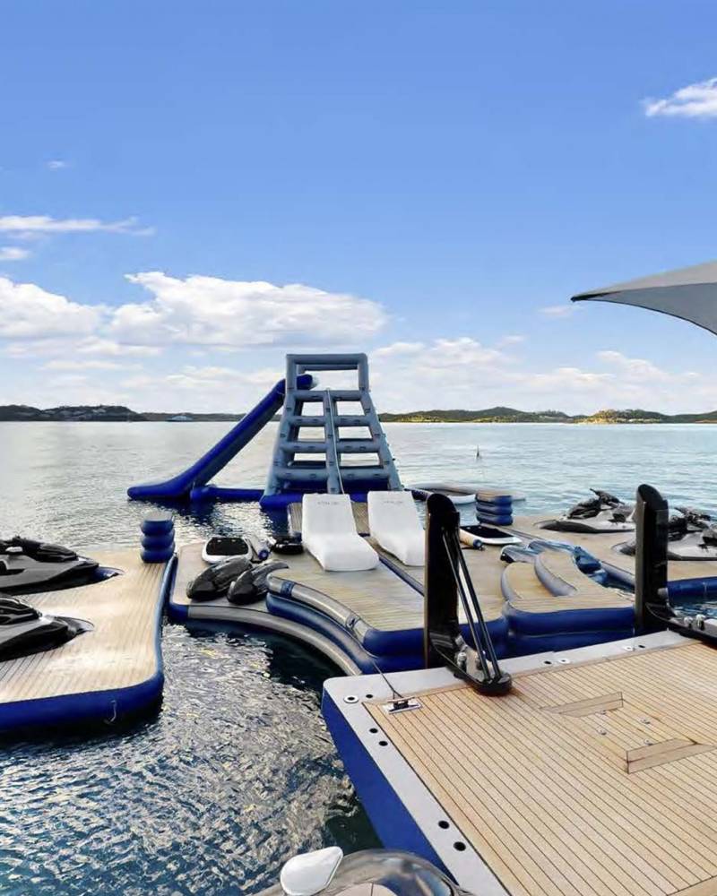 A floating water park with jet skis, a slide, and lounge chairs awaits on a serene lake under a bright blue sky.
