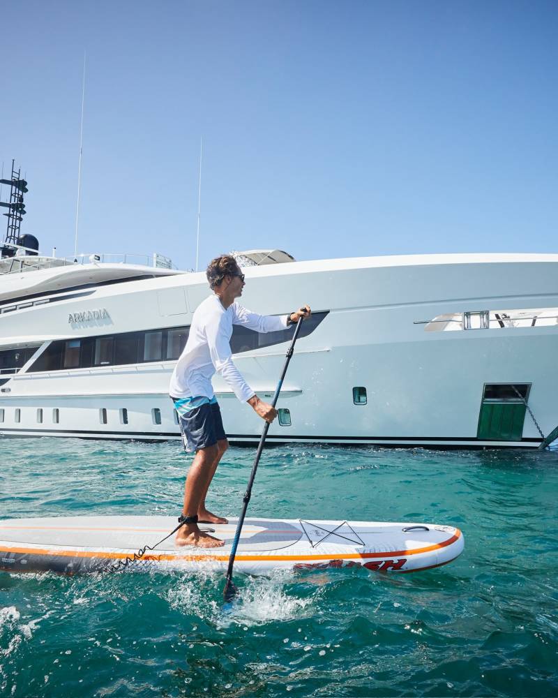 A man paddleboards on a calm sea with a luxurious yacht anchored in the background.