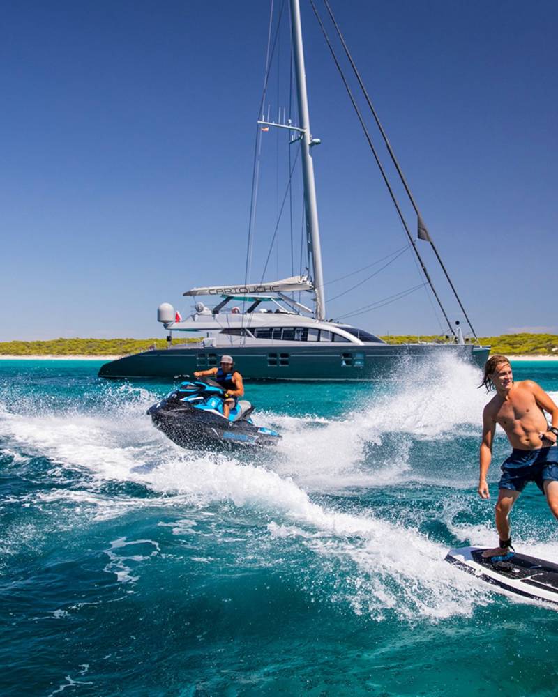 A man enjoys a thrilling ride on a jet ski while another surfs behind a large catamaran on a sunny day.