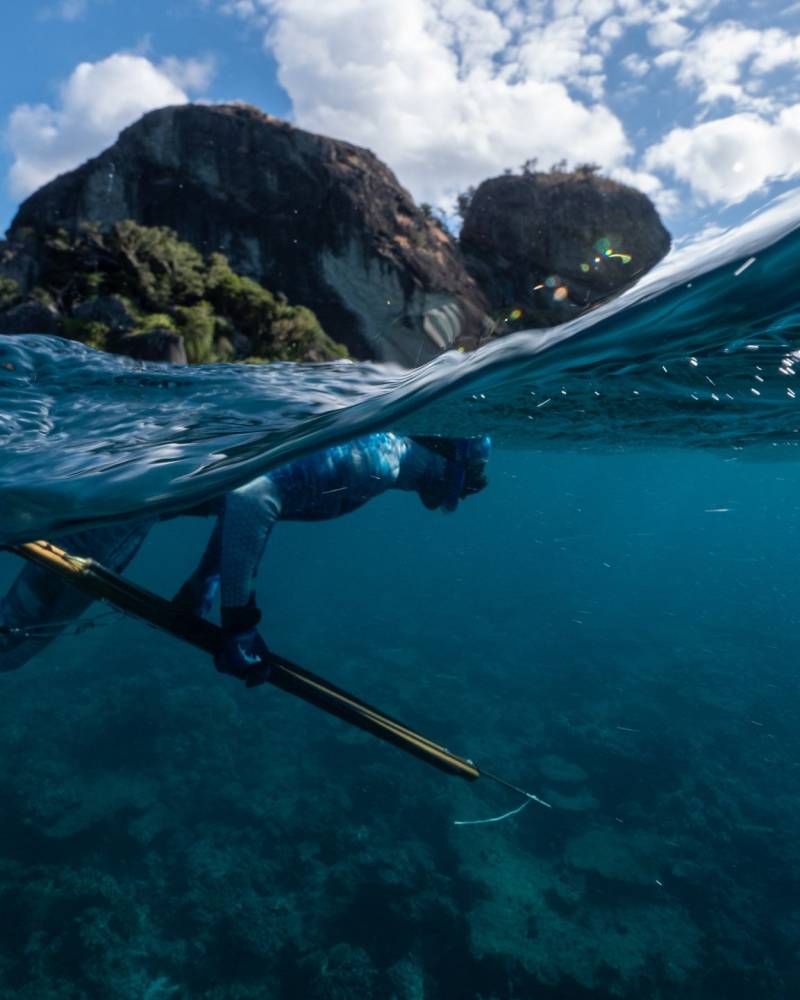 A spearfisher in a blue wetsuit glides through clear turquoise water near rocky islands under a bright blue sky.
